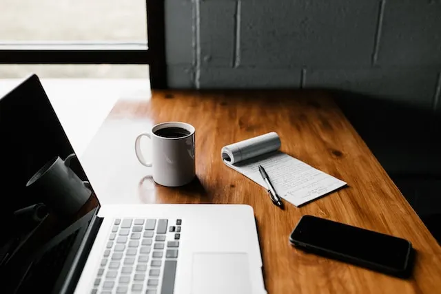 Picture of laptop, coffee, phone and pen and paper on desk.