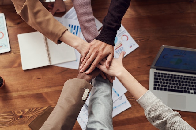 6 people putting there hands in together over the top of a desk.